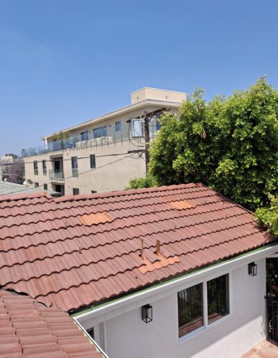 A red-tiled roof of a single-story house is seen in the foreground, with a modern multi-story building in the background under a clear blue sky.